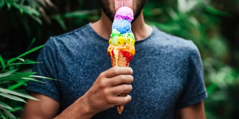 Um jovem tomando um sorvete nas cores do arco-íres (lgbtq+), o sorteve esta derretendo um pouco, ao fundo é possivel ver plantas verdes.